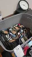Gray plastic basket filled with many Magic: The Gathering cards showing backs of cards with game logo, on floor near clock.