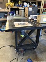 Rolling black table with caster wheels shown in garage environment, wooden insert where saw blade fits.