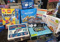 Photo showing all toy kits and educational workbooks arranged on a table. Includes LEGO Gadgets, Steel Works motorcycle kit, NextMaker coding box, Giggleway electric motor robot kit, Fraction Formula game, chemistry experiment book, Scratch coding workbook, and Silbird solar robot kit.