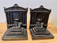 Pair of bronze bookends side by side showing classical columns, decorative pediment with rosette, and oil lamp on books in front