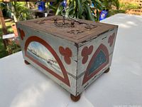 Angled view of rectangular jewelry box with painted landscape scenes on side panels and red arch decorative details. Key inserted in lock on lid, iron handle on top, set outdoors on white surface.
