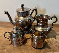 Photo showing the full antique silverplate tea set from a frontal angle on a wooden surface, including teapot, sugar bowl, water pitcher, and creamer with visible etched floral designs and ornate handles.