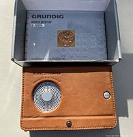 Top view of the Grundig G-2000A radio in brown leather case with speaker and power button, laid next to its original box showing brand and design by Porsche.