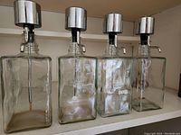 Four clear rectangular glass liquor bottles with metal bar top dispensers arranged side by side.