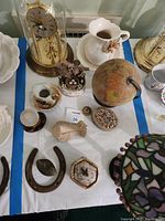 Top-view photo showing a variety of decorative items on a white surface with blue tape borders. Items include a glass dome clock, ceramic pitcher, globe, horseshoe, trinket boxes, and small collectibles.