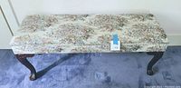 Front view of the antique storage bench with floral fabric and cabriole legs on blue carpet.