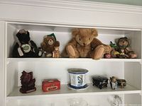 Photo of a white shelving unit showing a top and second shelf with various collectible and decorative items. Top shelf contains plush teddy bears in assorted clothing. Second shelf contains Asian inspired decor including red samurai statue, lacquered box, blue and white ceramic footed base, black footed stands, and small carved buddha figure.
