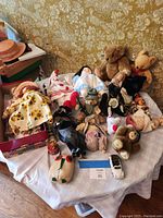 Photo showing a group of vintage dolls, teddy bears, plush animals, tin toy, and a small die-cast car arranged on a white tablecloth on a round table.