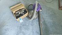 Full photo showing Dyson DC39 vacuum cleaner laid on carpet with hose and wand, and a cardboard box containing multiple vacuum attachments.