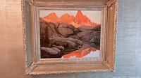 Full view of the framed landscape painting showing orange-hued mountain peaks, rocky foreground, and reflection in water.