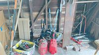 Photo showing miscellaneous garden tools and equipment inside a garage including red fuel containers, loppers, shovels, rakes, and a box with assorted hand tools