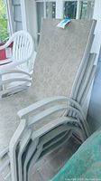 Stack of four white metal framed outdoor patio chairs with beige floral patterned cushions on seats and backs, dust visible, stored under covered porch.