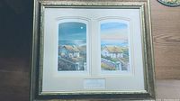 Framed artwork showing two prints of rural cottages and landscapes under serene sky in subtle colors, separated by matting and surrounded by gold textured frame.