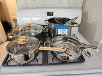 Photo of stainless steel pots and pans with gold accents arranged on a white stove, showing three pots (two with glass lids) and a frying pan, each with matching spoons