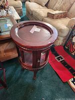 Full view of round wooden side table with detachable tray top and glass door cabinet below.