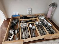 Top-down photo showing collection of silverware organized in wooden trays, includes multiple forks, spoons, knives, and serving utensils all with decorative handles, some in original boxes.