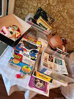 General assortment of vintage toys and costumes arranged on table with Batman/Superman costume box, mask, and art supplies visible.