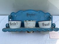 Front view of blue enamel wall shelf rack with three enamel pots labeled 'Seife', 'Streu', and 'Sand'. Shows shelf design and placement of pots.