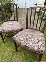Pair of antique wooden chairs with leopard print upholstered seats shown outdoors on grass. Both chairs have vertical slat backs and leopard print fabric seats.