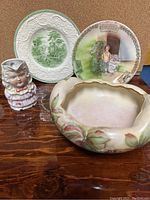 Four porcelain items arranged on wooden surface: hand painted floral bowl, decorative plate with child and lamb, green and white embossed plate, and small face jug