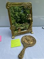 Two brass mirrors displayed on a table outdoors. Larger rectangular standing mirror with floral and figurative decoration. Smaller hand mirror with embossed figure design.