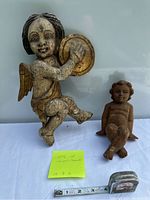 Two wooden carved cherubs placed on a white background, a tape measure for scale showing 12 inches, one larger painted cherub holding a tambourine and a smaller unpainted cherub sitting.