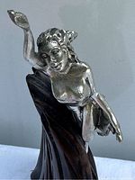 Front view of metal female figurine mounted on carved wooden base, showing detailed facial features and raised arm.