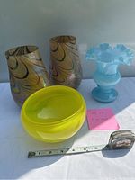 Four colorful glass vases and a glass bowl displayed on a white surface with measuring tape showing size