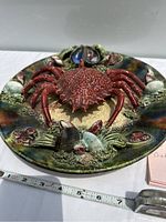 Front angle view showing 3D red crab and colorful surrounding sea life motifs on the plate surface, with a tape measure for size reference.