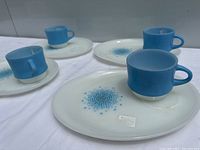Four sets of round white snack plates with blue starburst pattern each paired with a blue glass cup, showing surface wear and vintage condition.