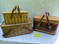Image showing three old metal picnic baskets; two yellow brown woven-effect metal baskets with wooden handles stacked on left and one tartan plaid metal basket with red folding handles on right on a white cloth background.