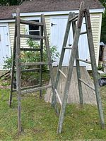 Two old wooden farm ladders standing outdoors on grass in front of a shed, showing full ladder height and structure with aged wood and chipping green paint.