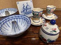 Overall view of Quimper porcelain items including egg cups, honey pot, bunt pans, and tile on wooden surface.