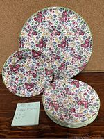 Full view of one large cake plate (11 inch diameter) behind six smaller dessert plates (7 inch diameter) with dense multicolored floral pattern and gold trim.