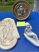 Photo of all three items together, showing the white ceramic nude figurine seated in front of the two wall plaques (one rectangular with a woman figure in relief, one round with floral motif)