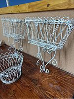 Two white vintage wire wall-mounted planters with curly brackets and lattice design, shown from angle on wooden table against corkboard wall, alongside matching wire pot holder basket