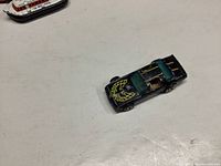 Black 1977 Hot Wheels Hot Bird die-cast toy car with yellow flame decals on the hood and teal-tinted windows on a white surface.