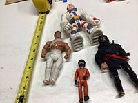 Four assorted vintage style plastic action figures on a white surface next to a measuring tape showing about 6 inches for the tallest figure.