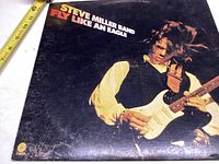 Front cover of Steve Miller Band's Fly Like An Eagle album showing Steve Miller playing guitar, measuring tape placed for scale.