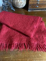 Close-up of folded cherry red mohair throw showing soft texture and fringed edge detail, placed on wooden surface with drawer and decorative items in background.
