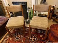 Two teak framed mid-century chairs with oatmeal textured fabric upholstery placed on a patterned rug near a fireplace and decorative items in the background.