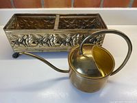 Photo showing the brass rectangular garden pot with intricate floral repoussé design beside the brass watering can to illustrate both items in the set.