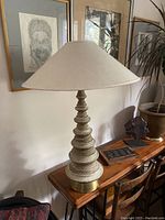 Full view of the lamp placed on a wooden console with framed art background, showing the overall height and profile of the lamp with its large white tapered shade and stacked disc base.
