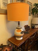 Full view of the lamp showing the cream chalkware base with tangerine and chocolate circles and stripes, topped with a tangerine fabric shade on wooden console.