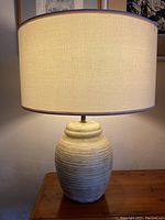 Full view of mid-century ceramic table lamp with wide cylindrical beige fabric shade on wooden table.