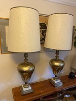 Pair of vintage gold floral motif table lamps on wooden surface against wall art background with cream cylindrical lampshades.