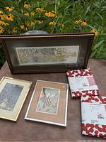 Photo showing three framed art prints and two unopened Gullbergsö pillow covers on a wooden table outdoors with flowers in the background.