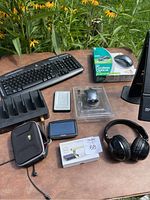 Lot contents displayed on a wooden table outdoors, including Logitech mouse in packaging, keyboard, headphones, charging dock, webcam, external hard drive, and carrying case.