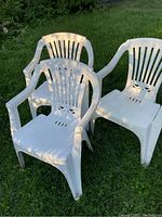 Three white plastic lawn chairs arranged on grass with slatted backs and integrated armrests.