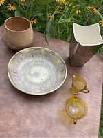 Five pottery items on table outdoors including tan bowl, large swirl glazed plate, tall brown and white vase, and two small amber glass pitchers.